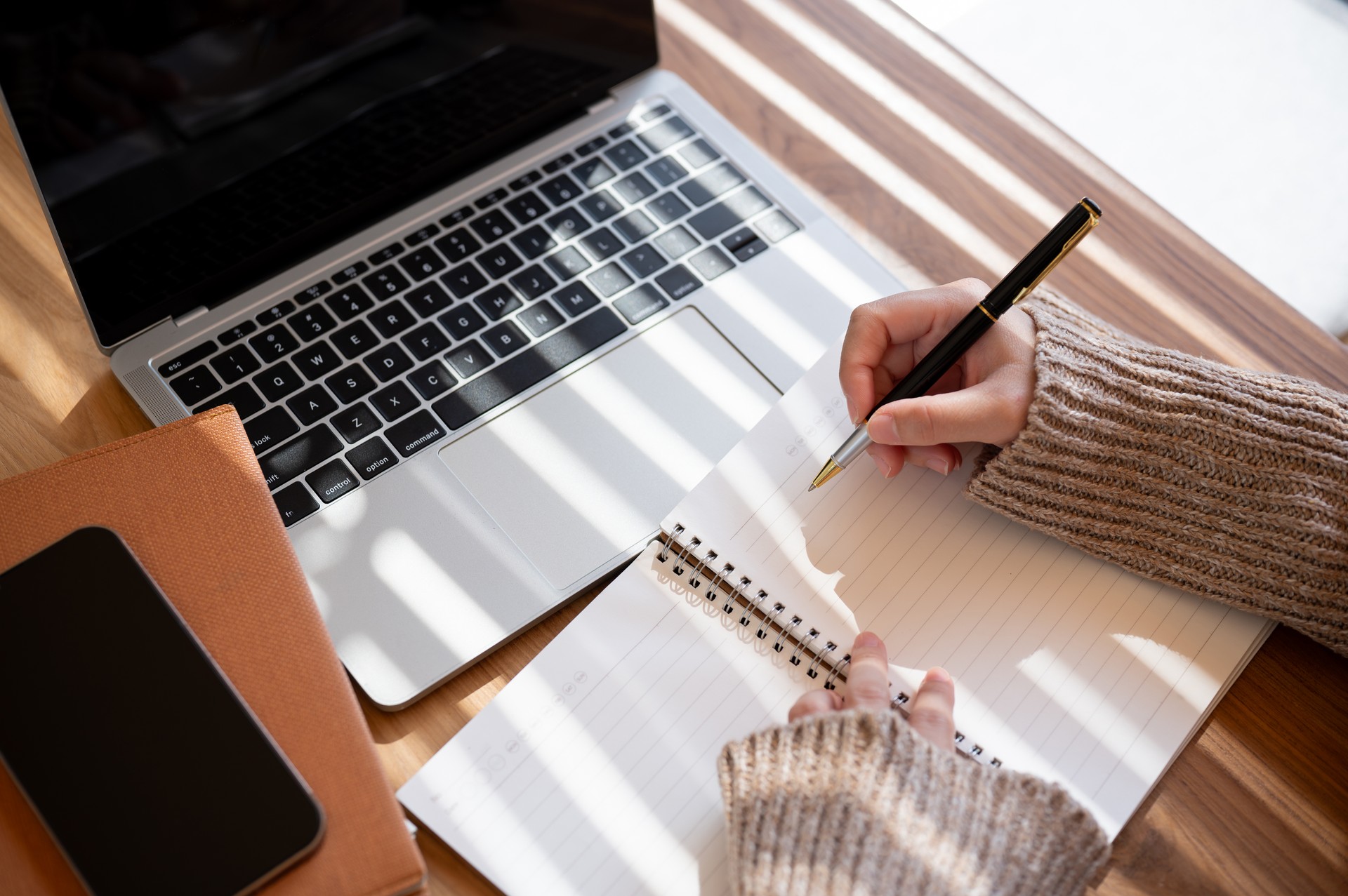 A top-view image of a woman is taking notes in her notebook while working on her laptop indoors.